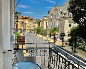 A balcony with a wicker chair and a small table, overlooking a street with buildings and palm trees. - Villa Sokol - Belle Epoque studio (Opatija)