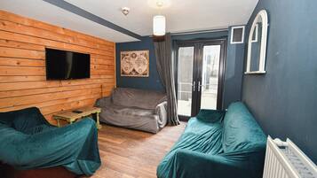 A room with a wooden wall, a flat-screen TV, a sofa, a chair, and a window with curtains.