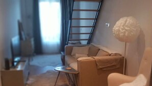 En moderne stue med sofa, stol, lille bord og gulvlampe. En trappe er synlig i baggrunden.
