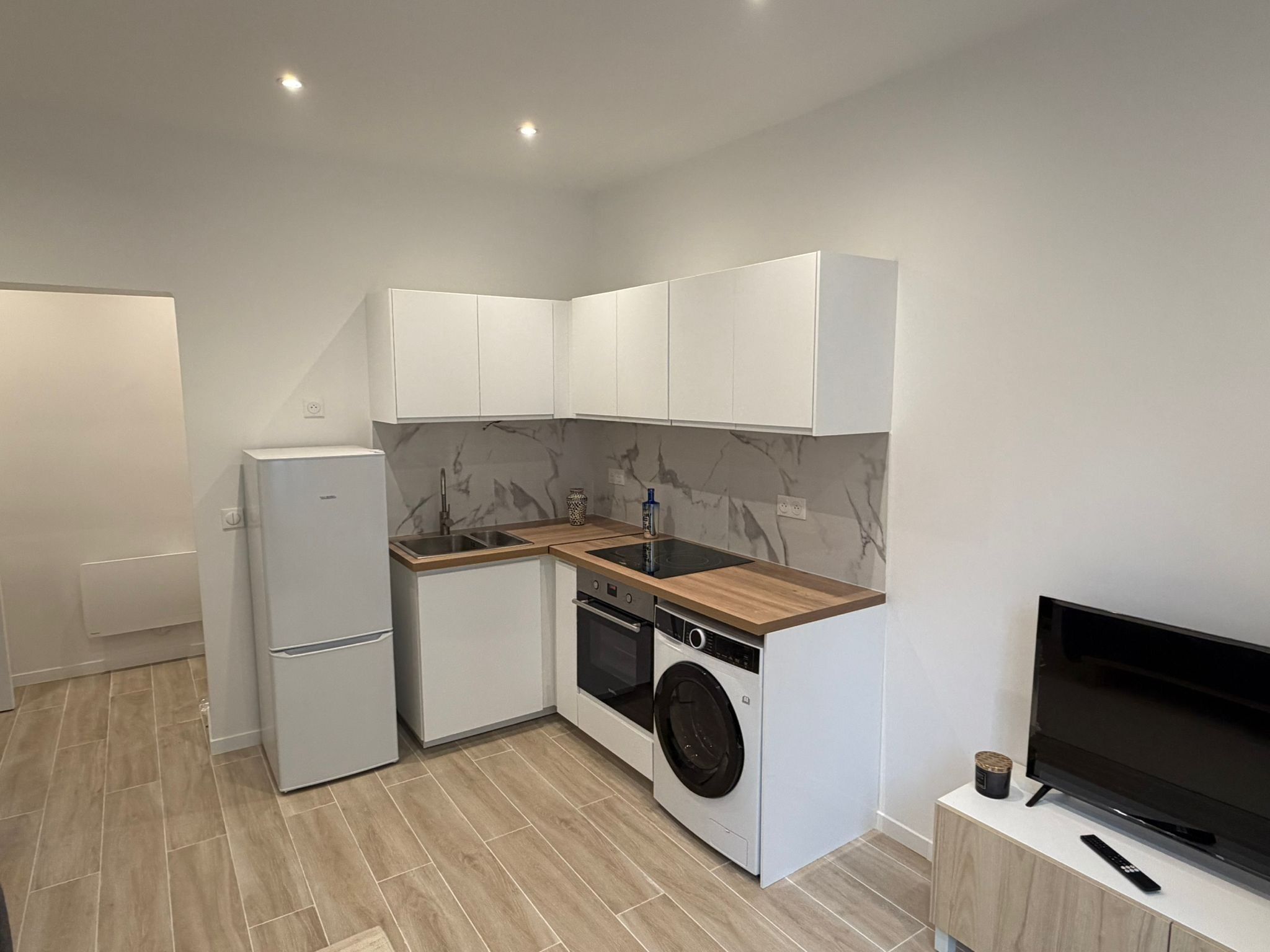 Eine moderne Küche mit weißen Schränken, einem Einbauofen, einer Waschmaschine und einem Flachbildfernseher auf einem Holzboden.