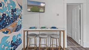 Een eethoek met een aan de muur bevestigde televisie, een tafel met krukken, en een muurschildering.