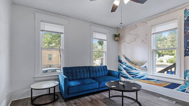 Een woonkamer met een blauwe bank, twee ronde tafels, een plafondventilator en een groot raam met uitzicht.
