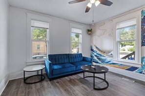Een woonkamer met een blauwe bank, twee ronde tafels, een plafondventilator en een groot raam met uitzicht.