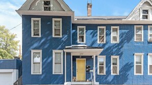 Een blauw huis met twee verdiepingen, een gele voordeur, huisnummer 677, en een schoorsteen.