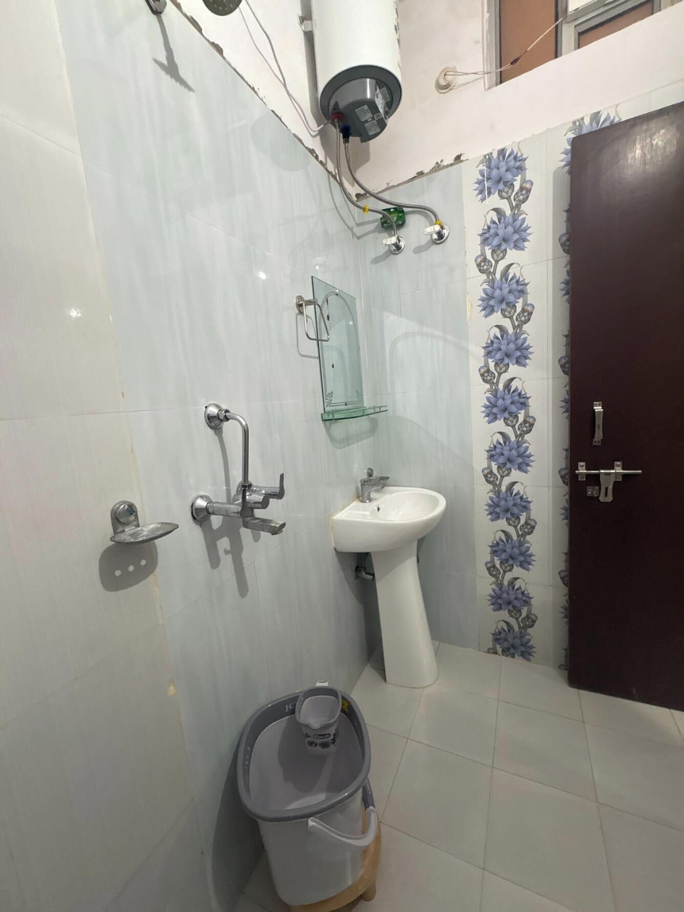 A bathroom with a toilet, bidet, sink, and shower. There is a bucket on the floor and a door with a handle.