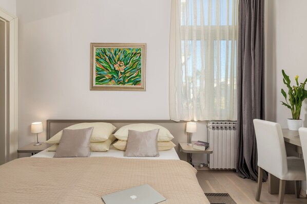 A hotel room with a bed, bedside lamps, a desk, a potted plant, and a painting on the wall.