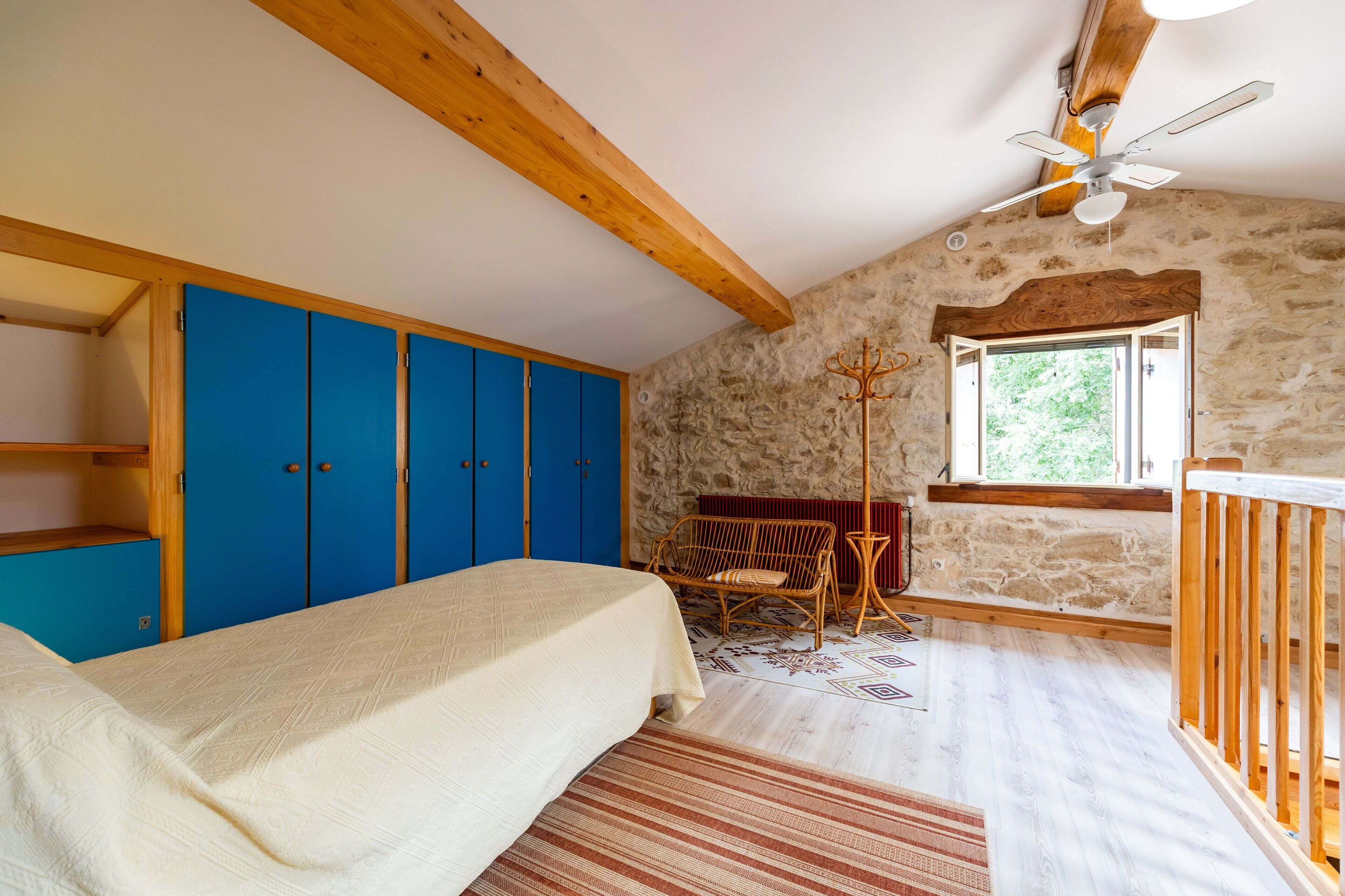 Un dormitorio con cama, armario azul, paredes de piedra, techo de madera y ventana.