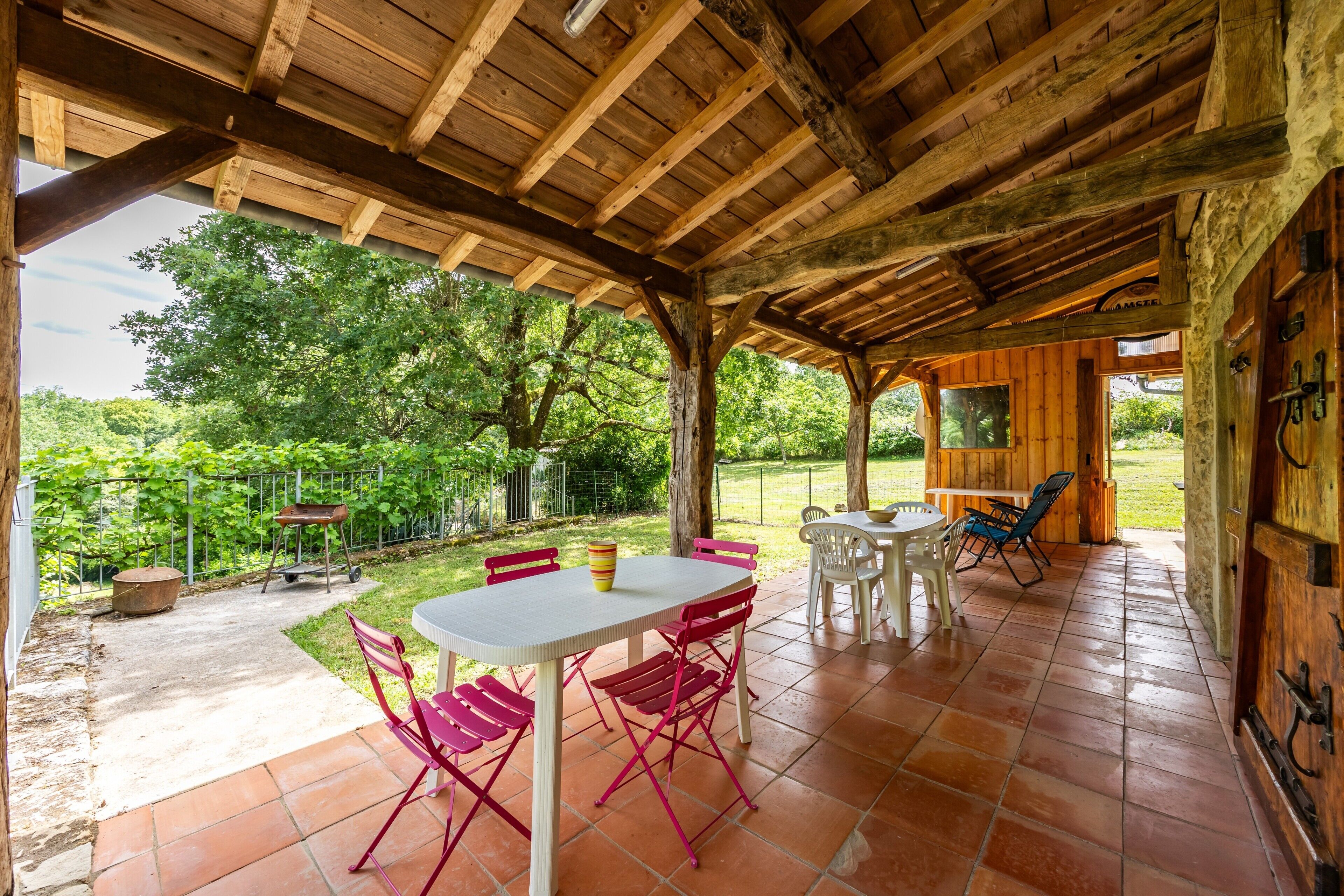 Une terrasse extérieure avec des poutres en bois, une table et des chaises, et une vue sur un jardin.