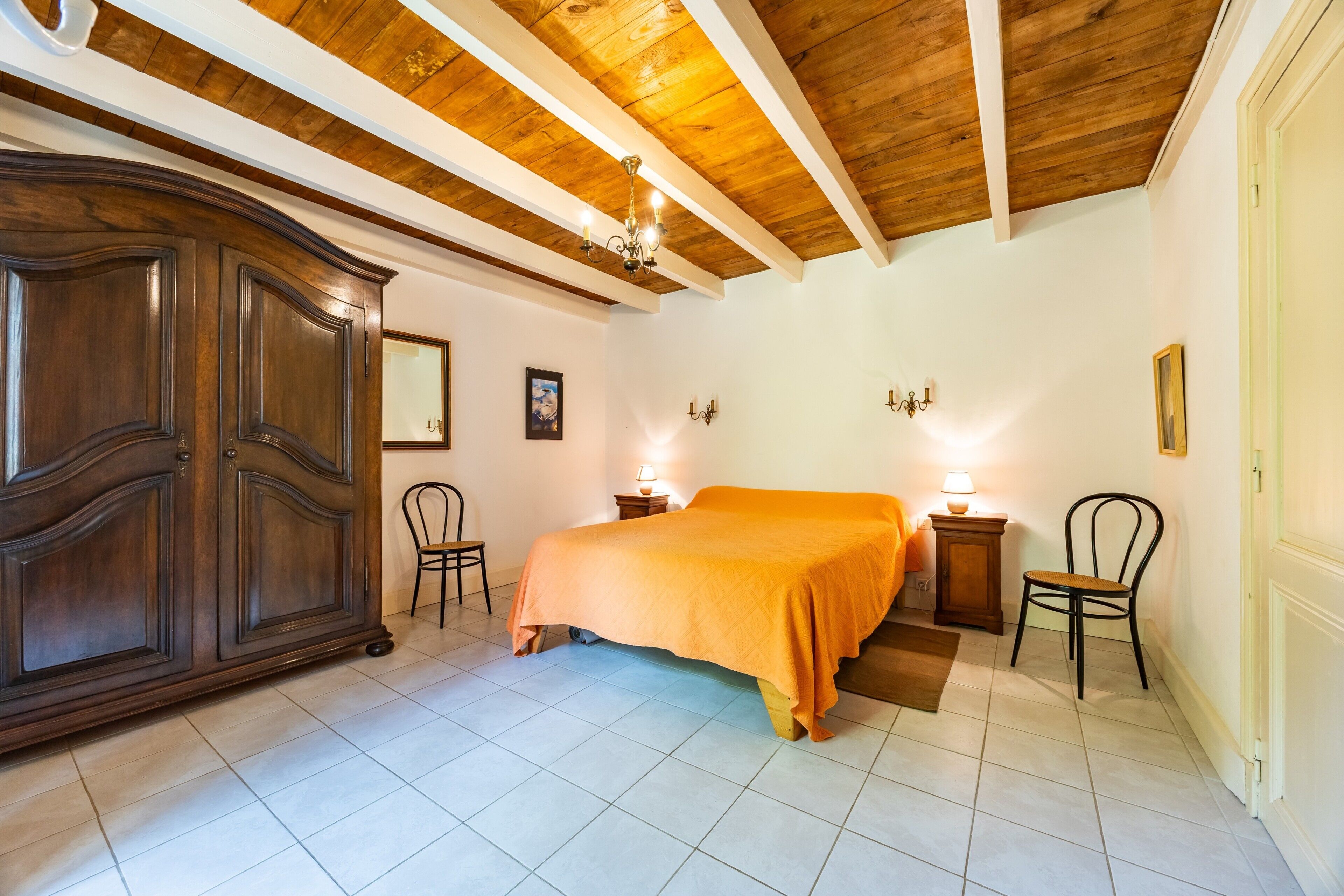 A bedroom with a bed, wooden wardrobe, two chairs, a nightstand, and a chandelier.