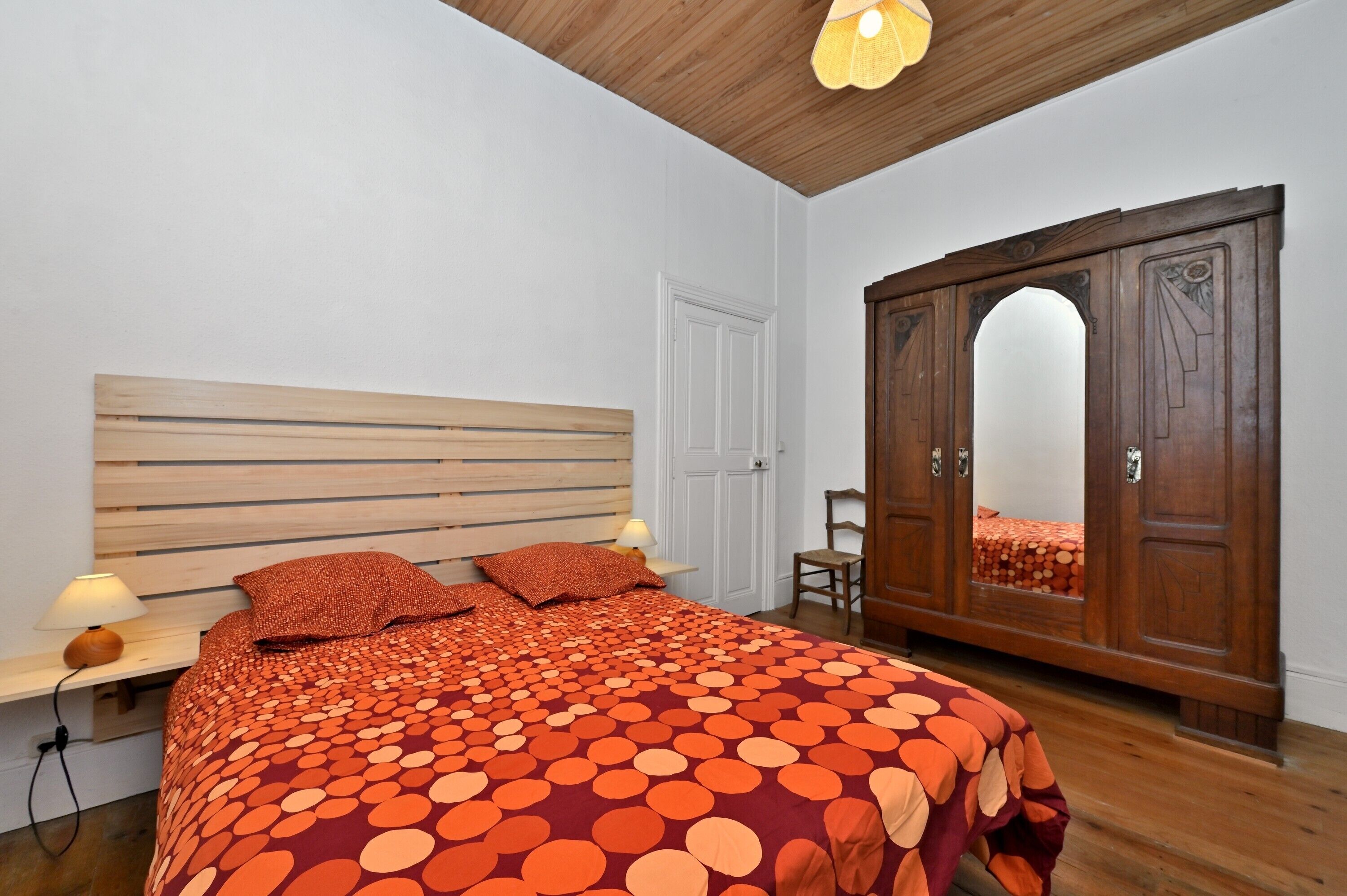 A bedroom with a wooden headboard, a large bed with a polka dot blanket, a wooden wardrobe, and a mirror.