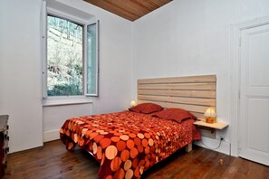 Une chambre à coucher avec une tête de lit en bois, un lit recouvert d’une couvre-lit à motifs, une table de chevet avec une lampe et une fenêtre donnant sur des arbres.