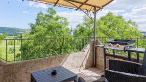 Un espace extérieur abrité, doté d’un barbecue, de meubles en osier et offrant une vue sur la verdure et les collines lointaines.