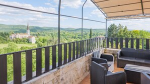 A covered outdoor balcony with wicker furniture, a stone wall, and a view of a church and green landscape. - Les Traverses Gîte with Shared Pool, Wi-Fi, and Air Conditioning (Chauzon)