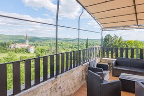 A covered outdoor balcony with wicker furniture, a stone wall, and a view of a church and green landscape. - Gîte “Les Traverses” with Shared Pool, Wi-Fi, and Air Conditioning (Chauzon)