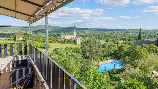 A balcony with a view of a church, a swimming pool, and surrounding greenery. - Les Traverses Gîte with Shared Pool, Wi-Fi, and Air Conditioning (Chauzon)