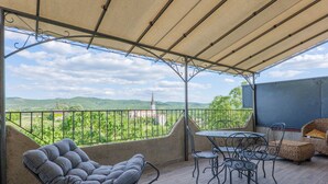 A covered patio with a table and chairs, a cushioned chair, and a view of green hills and a distant tower. - Gîte "Le Barquetier" with Shared Pool, Wi-Fi, and Air Conditioning (Chauzon)