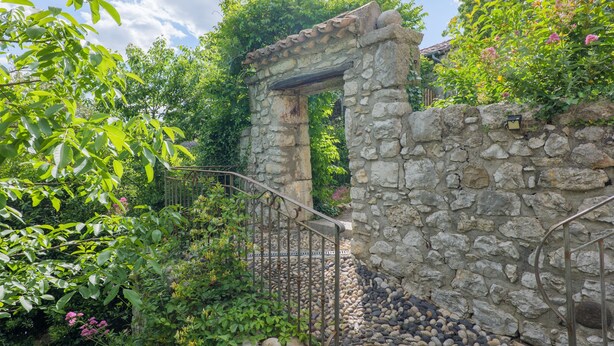 A stone archway leading to a garden area with a metal gate, surrounded by lush greenery and flowering plants. - Gîte "Le Barquetier" with Shared Pool, Wi-Fi, and Air Conditioning (Chauzon)