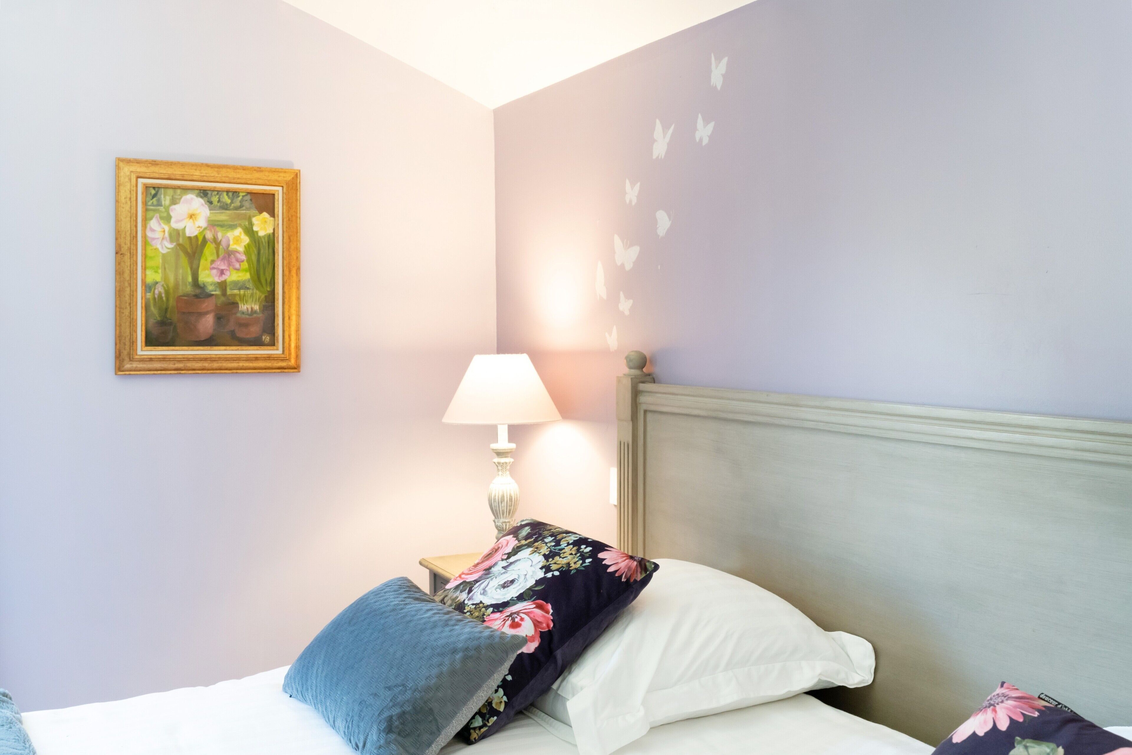 Ein Schlafzimmer mit einem Bett, Kissen, einem Nachttisch mit Lampe, einem floralen Gemälde und Schmetterlingsmotiven an der Wand.