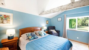 Un dormitorio con cama, mesita de noche, silla, ventana y un cuadro en la pared.