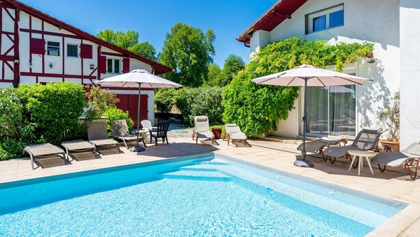 A swimming pool with lounge chairs and umbrellas, surrounded by a garden and two-story buildings. - Gite de la Barthe – near Dax (Garrey)