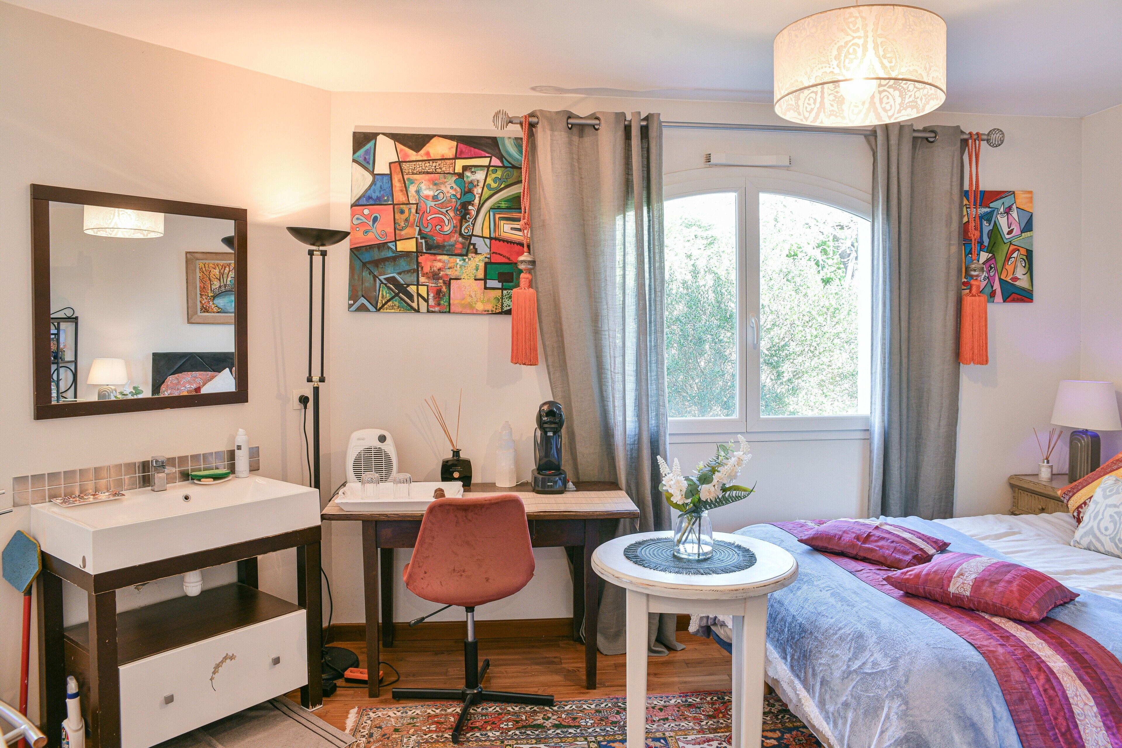 A bedroom with a bed, a desk, a chair, a mirror, a window with curtains, and a wall with abstract art.