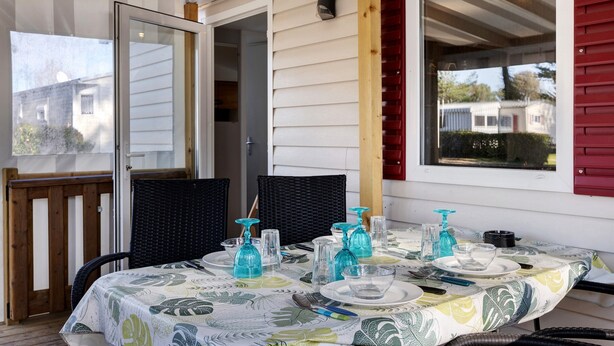 A covered patio with a table set for two, featuring clear glassware and a patterned tablecloth. - Mobile Home 148 in Bois Dormant Campsite, 2 Bedrooms, 4 Guests (Saint-Jean-de-Monts)