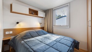 A bedroom with a bed, bedside table, wall-mounted shelf, and a window with curtains. - Mobile Home 148 in Bois Dormant Campsite, 2 Bedrooms, 4 Guests (Saint-Jean-de-Monts)