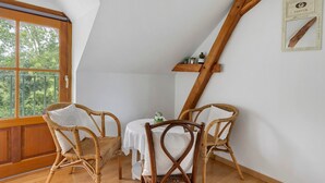 Een kamer met een schuine kap, een houten stoel, een klein tafeltje met een witte tafelkleed en een raam met uitzicht op groen.