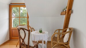 Een hoekzaal met een raam met houten kader, een ronde tafel met een witte tafelkleed en twee rieten stoelen.