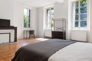 A bedroom with a bed, a desk, a chair, a radiator, and a window with a view of greenery.