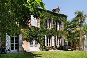 A building with a brick facade partially covered in ivy, white shutters, and a small garden area with a table and chair.