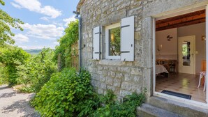 A stone building with a wooden door and window shutters, a visible interior with a dining area, and a view of greenery and hills in the background. - Gîte "Les Combes" with Shared Pool, Wi-Fi, and Air Conditioning (Chauzon)
