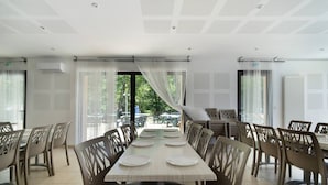 A dining area with a long table set for six, surrounded by chairs, with large windows and an air conditioning unit on the wall.