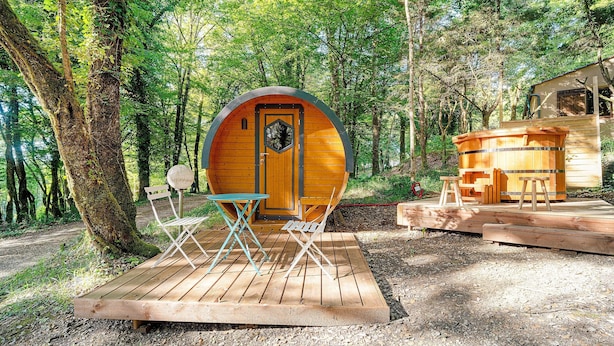 A wooden cabin with a circular door, surrounded by trees, and a small deck with folding chairs and a table.