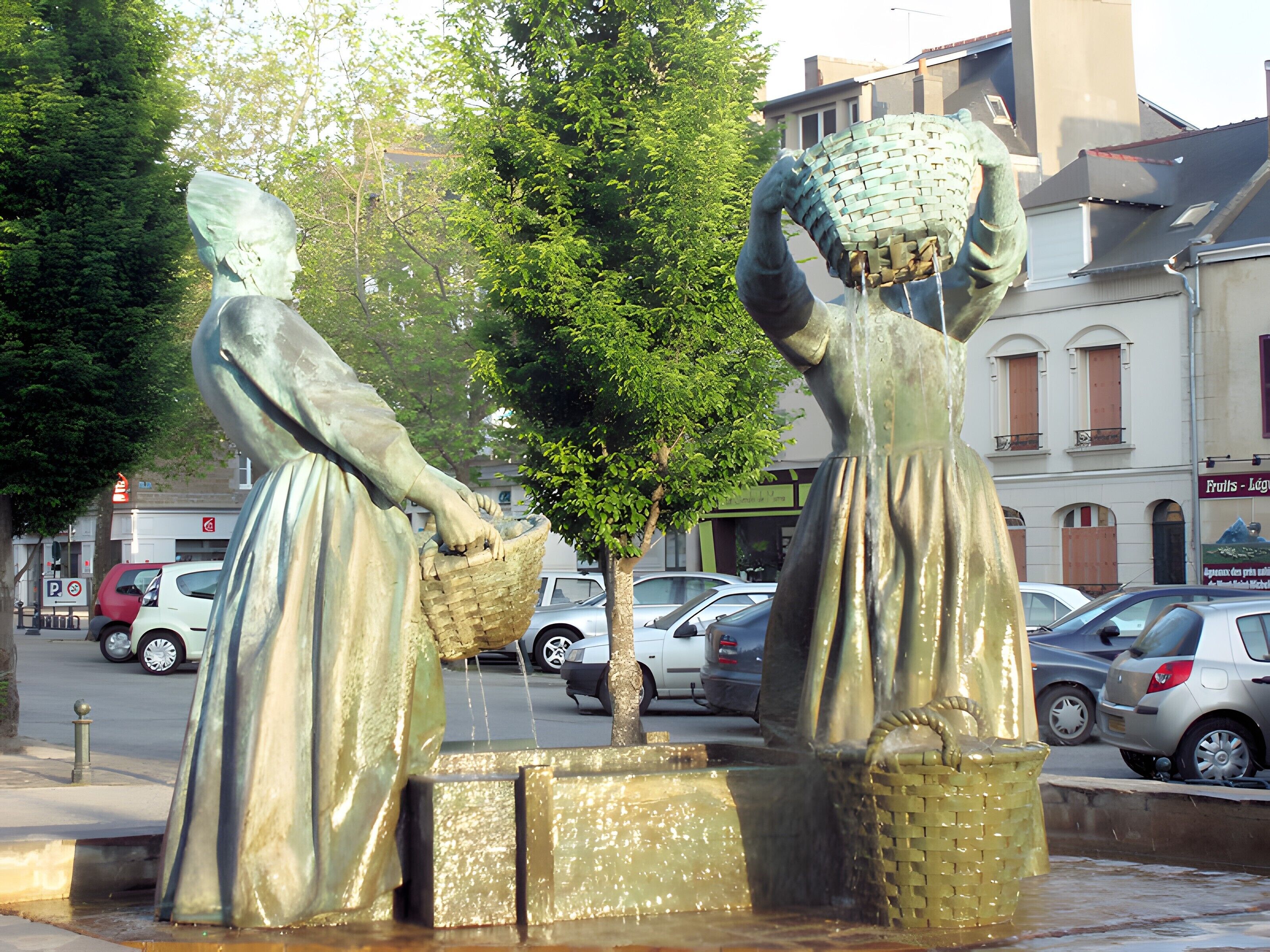 A statue of two figures carrying baskets, surrounded by a fountain, with cars and buildings in the background.