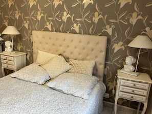 A bedroom with a tufted headboard, two bedside tables with lamps, and a floral wallpaper pattern. - Bed and Breakfast 'Chez Marie-christine - Les Fleurs' with Wi-Fi (Cancale)