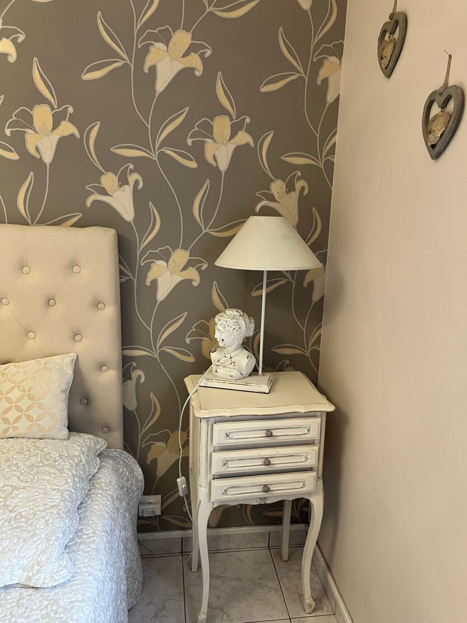 A bedroom with a tufted headboard, a bedside table with a lamp and a bust sculpture, and a patterned bedspread.