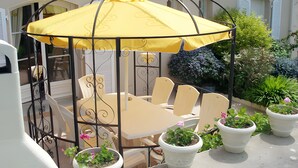 Un área de estar al aire libre con techo, con una mesa y sillas, rodeada de plantas en macetas.