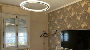 Un dormitorio con televisor grande, radiador, ventana con persianas y pared con papel tapiz floral.