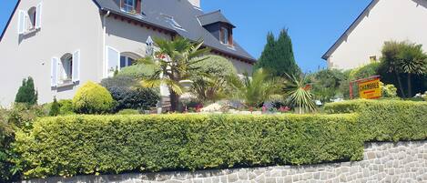 A two-story house with a stone wall in front, a well-maintained garden with various plants and flowers, and a sign indicating the name 'CHAMBRES.'