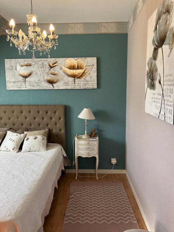 A bedroom with a tufted headboard, a white bedside table with drawers, a lamp, and a large floral artwork on the wall.