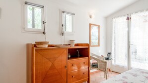 Un dormitorio con un aparador de madera, dos ventanas, una cama con colcha estampada y una silla de mimbre.