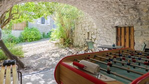 Une table de baby-foot à l’intérieur, dans une pièce aux murs de pierre, avec vue sur un jardin et une porte en bois.