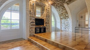 Une pièce aux murs de pierre, dotée d’une cheminée, d’un téléviseur et offrant une vue sur l’extérieur grâce à une grande fenêtre en arc de cercle.