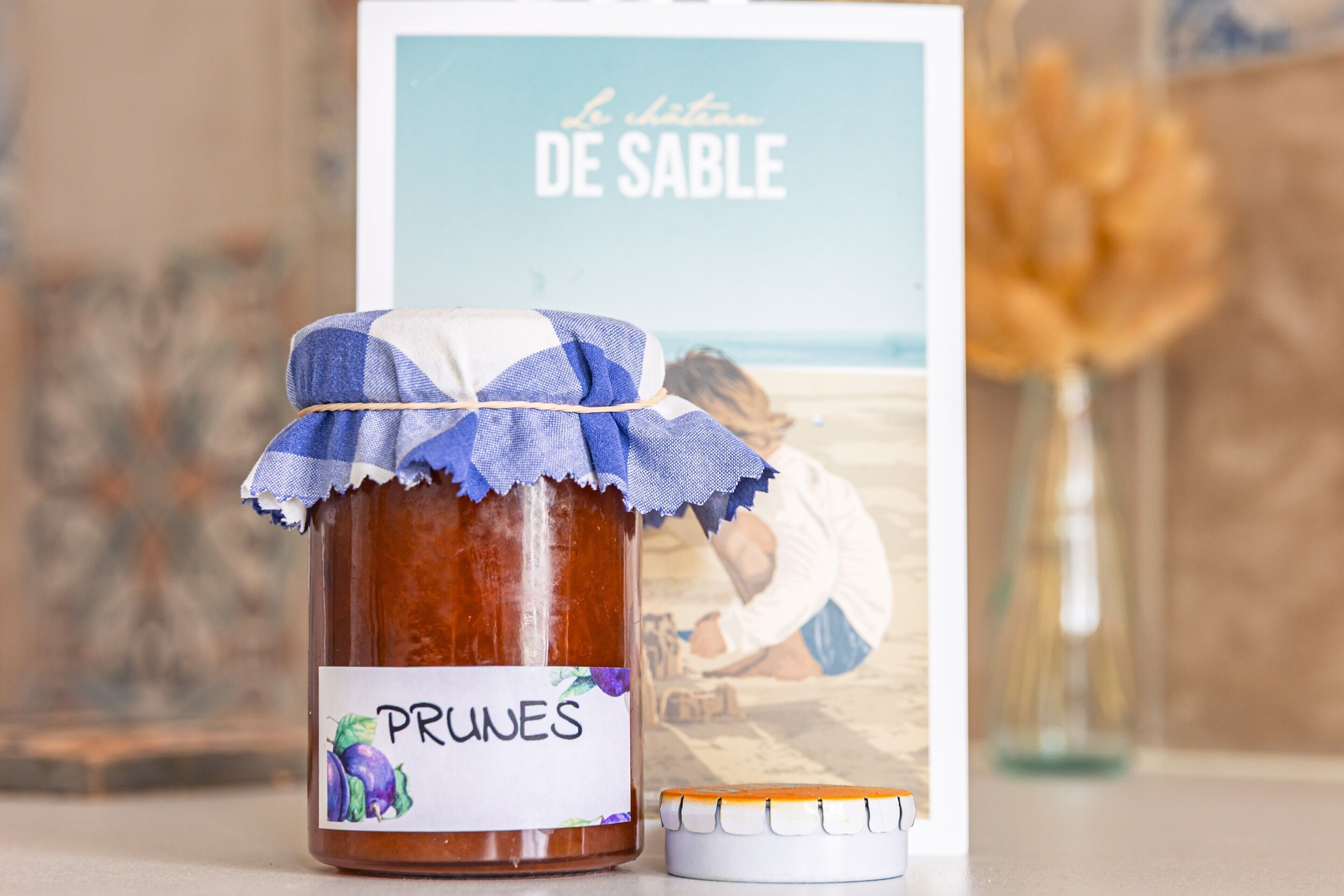A jar of prunes with a label, a small white container, and a framed picture in the background.