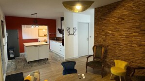 A modern living room with a kitchen in the background, a wooden accent wall, and a dining area with two chairs. - Gîte Les Pivoines with Private Terrace, Wi-Fi, and Air Conditioning (Saint-Robert)