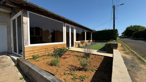A single-story building with large windows, a garden with various plants, and a paved driveway. - Gîte Les Pivoines with Private Terrace, Wi-Fi, and Air Conditioning (Saint-Robert)