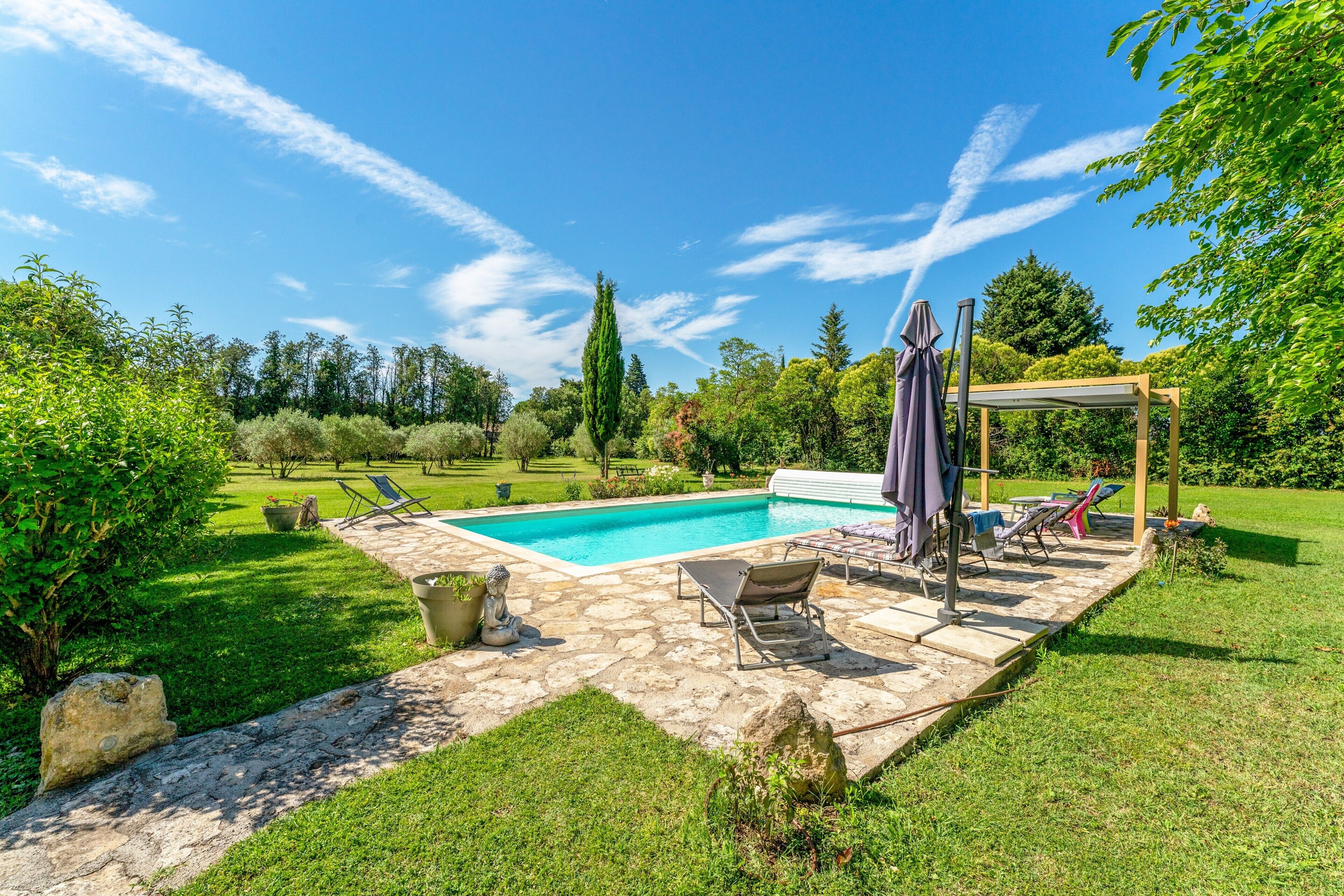Ein Garten mit Schwimmbad, Steinterrasse, Liegestühlen und einer Pergola mit Sonnenschirmen.