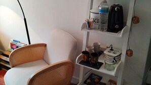 Ein Zimmer mit einem weißen Stuhl, einem weißen Regal mit verschiedenen Gegenständen und einer Wandablage mit einem Wasserkocher und einer Wasserflasche.