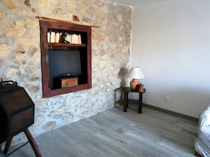 A stone wall with a television set in a wooden frame, a lamp on a small table, and a grill.